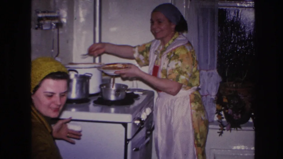 1966 Hoboken home-movie frame showing two women in a kitchen, with one serving food at a white stove and another smiling in a knit hat.