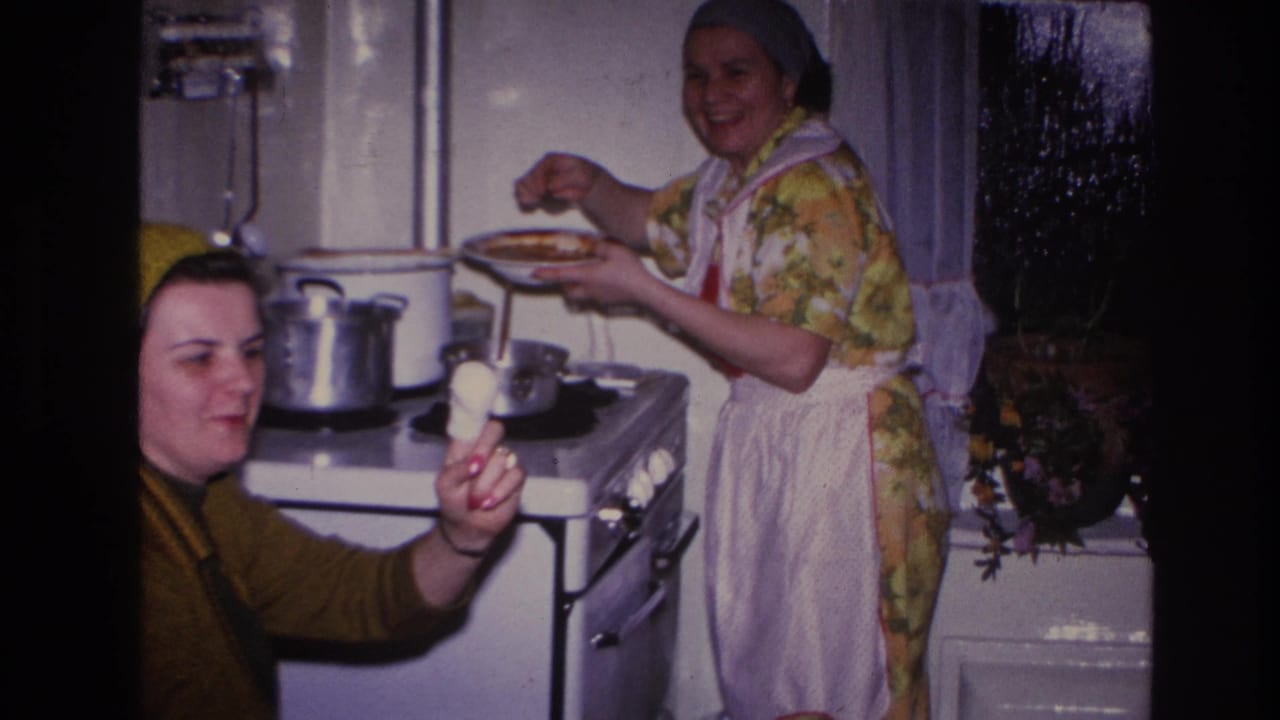 A seated woman holds up food on a spoon while another woman stands by the stove with a plate.