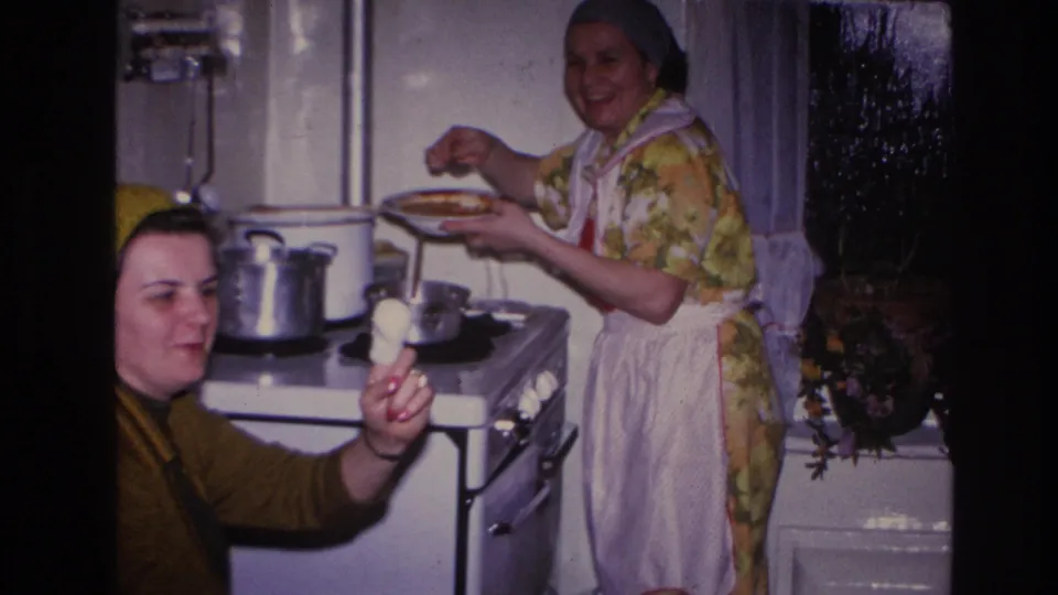 1966 Hoboken kitchen scene with a younger woman presenting a bite of food and an older woman serving beside cookware on the stove.
