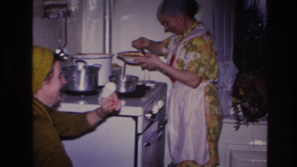 Candid 1966 Hoboken home-movie frame of two women laughing in a private kitchen as food is served from a pot on the stove.