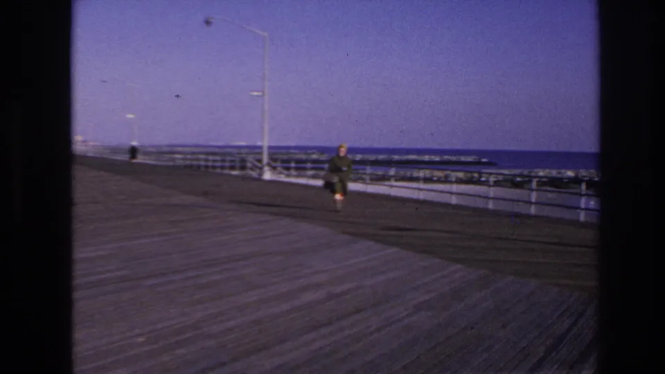 1966 Hoboken-area home-movie frame showing a distant person walking on a wooden boardwalk beside water and railings.