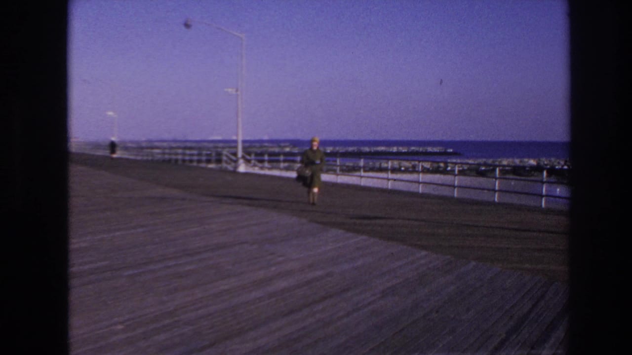 1966 Hoboken home-movie frame showing a woman with a hat and handbag walking along a waterfront boardwalk.