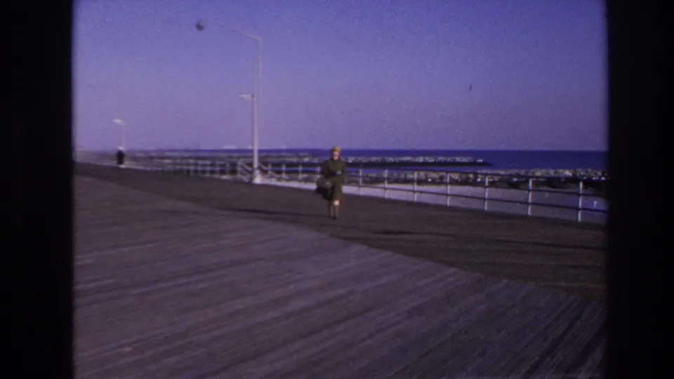 1966 Hoboken home-movie frame showing a woman with a hat and handbag walking along a waterfront boardwalk.