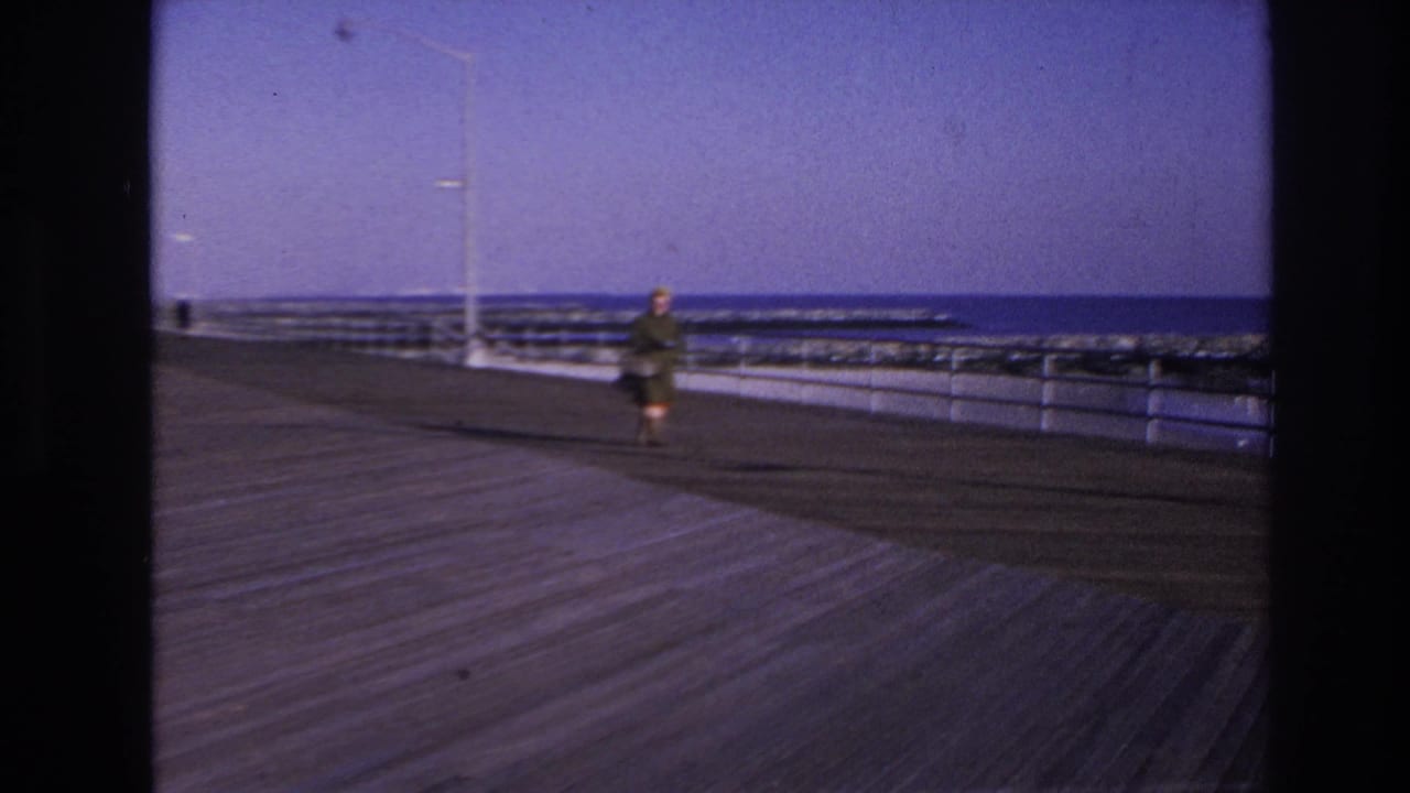 Woman on a boardwalk near a lamppost, with railings, waves, and horizon behind her.