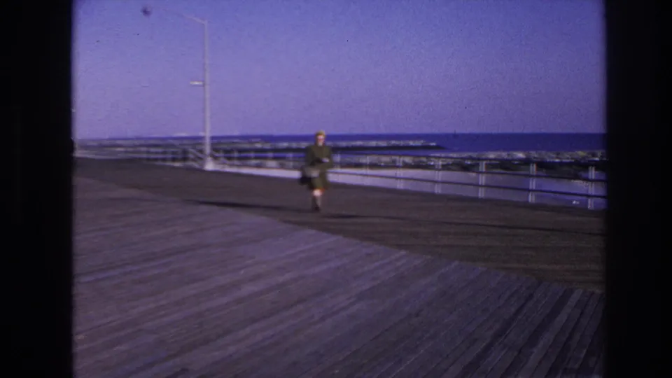 1966 Hoboken boardwalk frame showing a woman approaching the camera across sunlit planks by the waterfront.