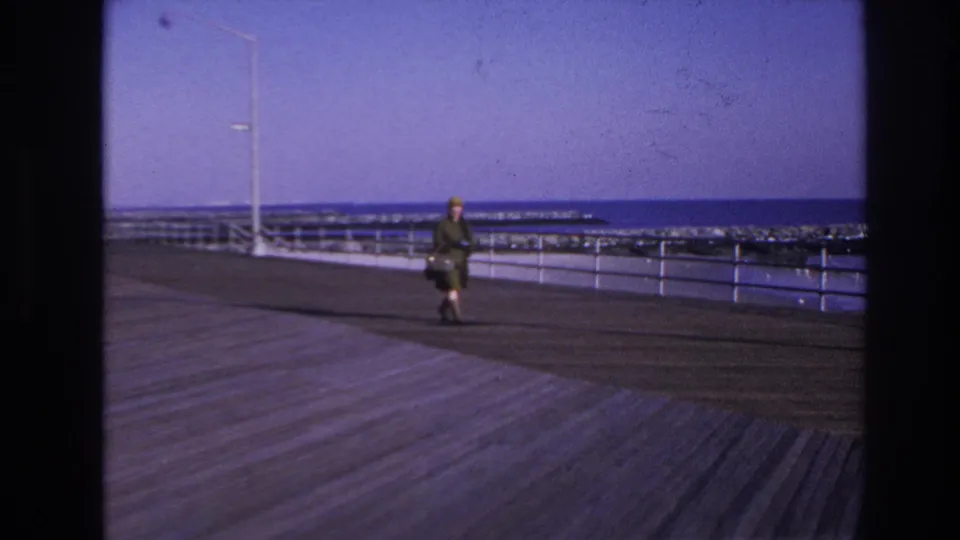 1966 Hoboken home-movie still with a woman in green-toned clothing carrying a handbag on the waterfront boardwalk.