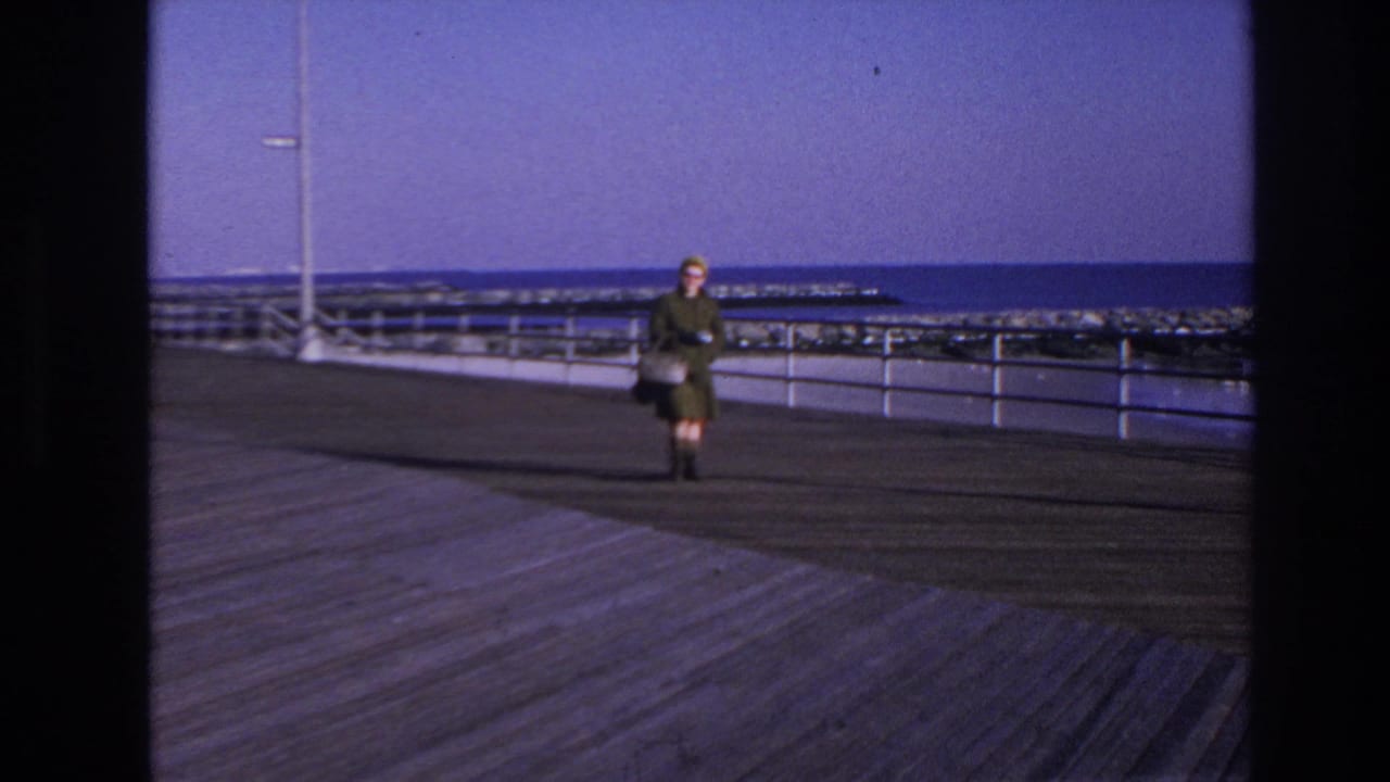 In 1966 Hoboken, New Jersey, a woman in a green coat and yellow hat walks toward the camera on a boardwalk with railing and water behind her.