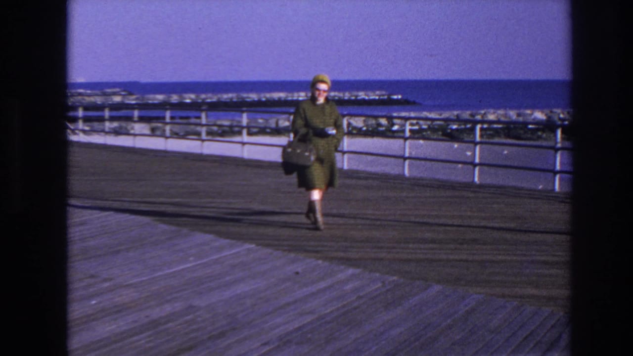 Woman carrying a dark handbag walks closer along the boardwalk near a white railing.