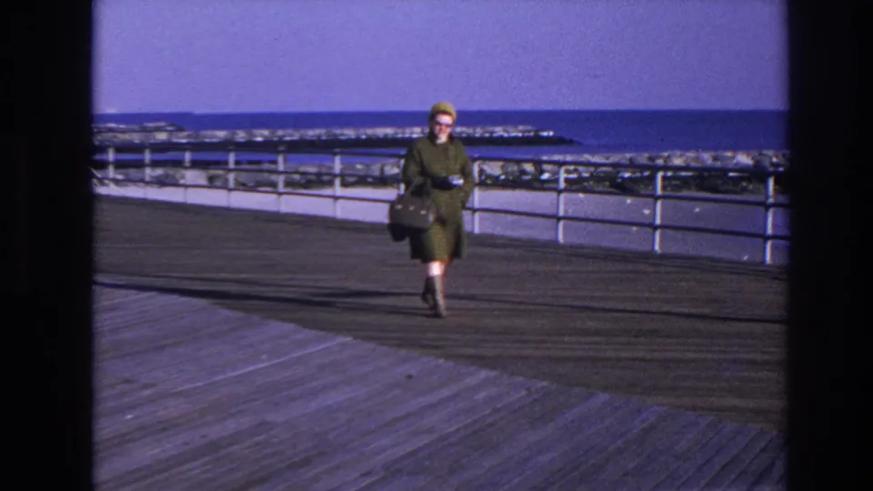 The woman in a green plaid coat, yellow hat, and sunglasses approaches on the Hoboken boardwalk, with water and rocks beyond the railing.