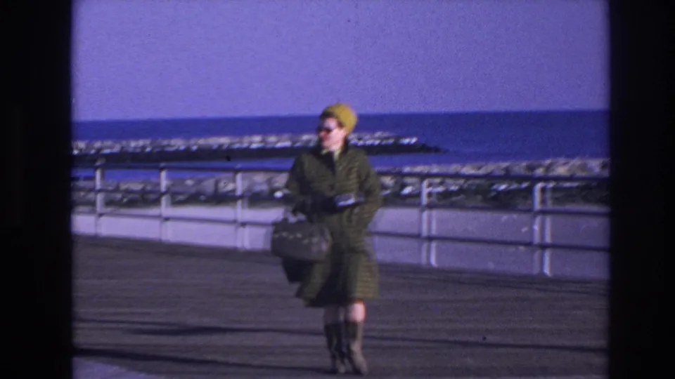 The 1966 boardwalk shot shows the woman closer to camera, wearing sunglasses and a yellow hat while holding a dark handbag near the waterfront railing.