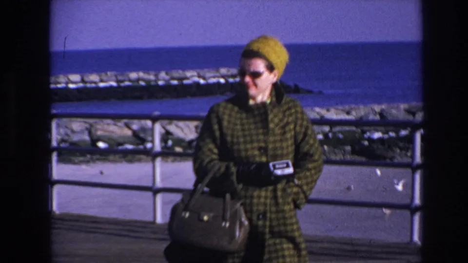 A closer 1966 Hoboken boardwalk frame shows the woman in a green plaid coat, yellow hat, sunglasses, and gloves beside the waterfront railing.