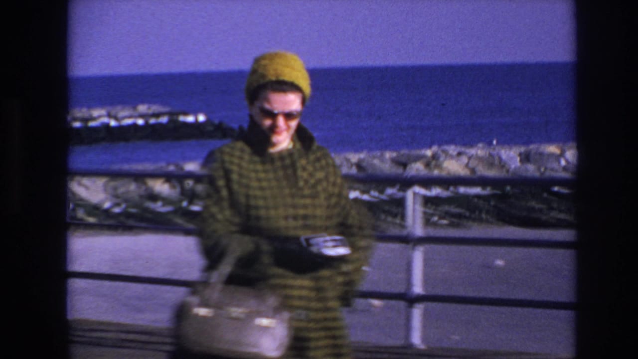 In 1966 Hoboken home-movie footage, a woman in a green plaid coat and yellow knit hat smiles near a boardwalk railing by the water.