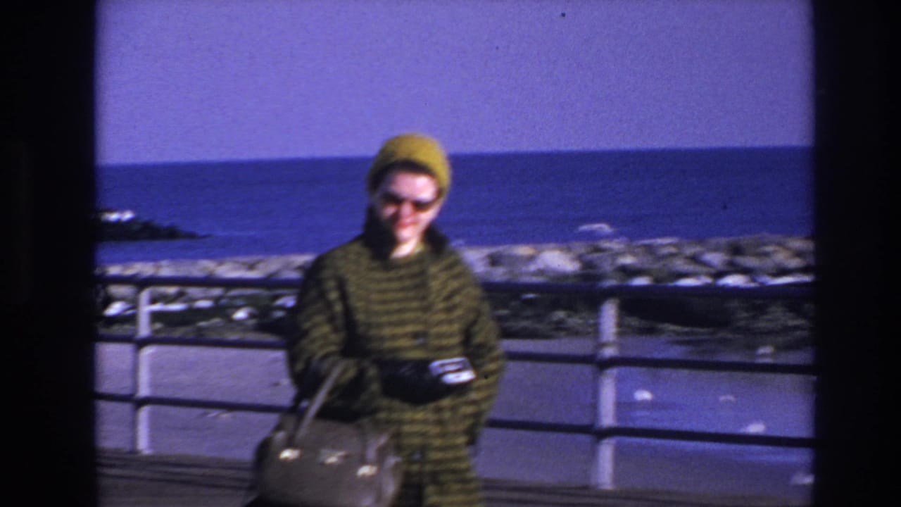 Woman walking along the boardwalk near a railing with rocks and blue water behind her.