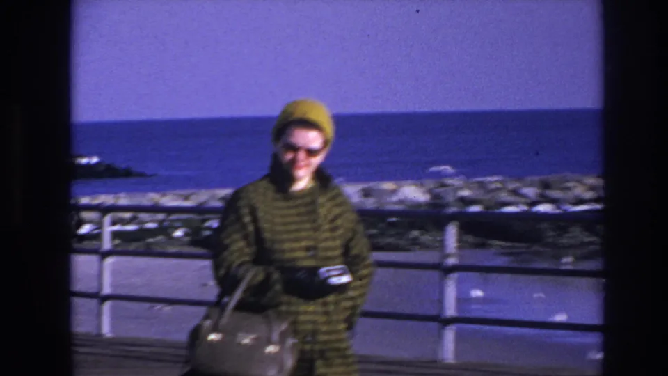 The woman continues along the Hoboken waterfront boardwalk, carrying a light handbag and holding a small camera near the railing.