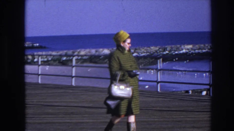 A side view shows the woman walking across the wooden boardwalk in Hoboken, with the waterfront railing and rocky edge behind her.