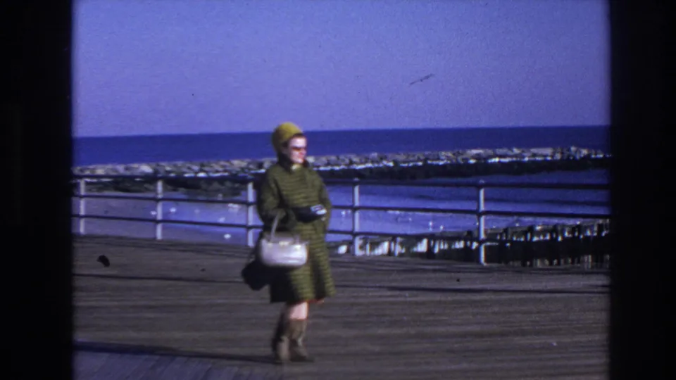 The woman appears farther from the camera as she walks along the Hoboken boardwalk, with the ocean and railing filling the background.