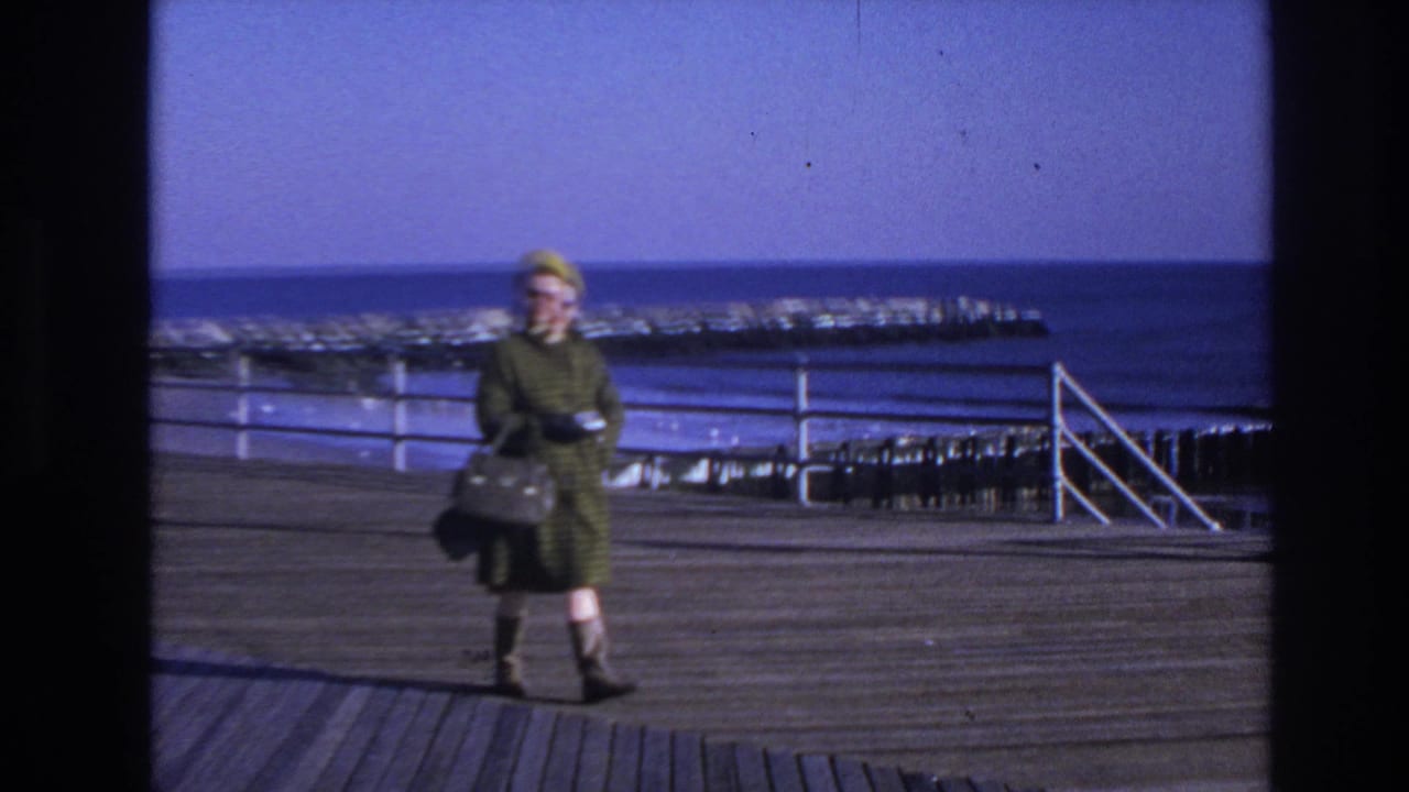1966 Hoboken home-movie frame showing a woman in a green coat and yellow hat walking on a wooden pier near choppy water.