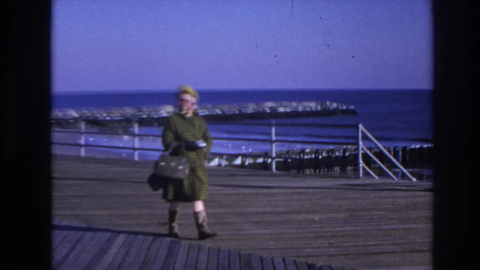 1966 Hoboken home-movie frame showing a woman in a green coat and yellow hat walking on a wooden pier near choppy water.