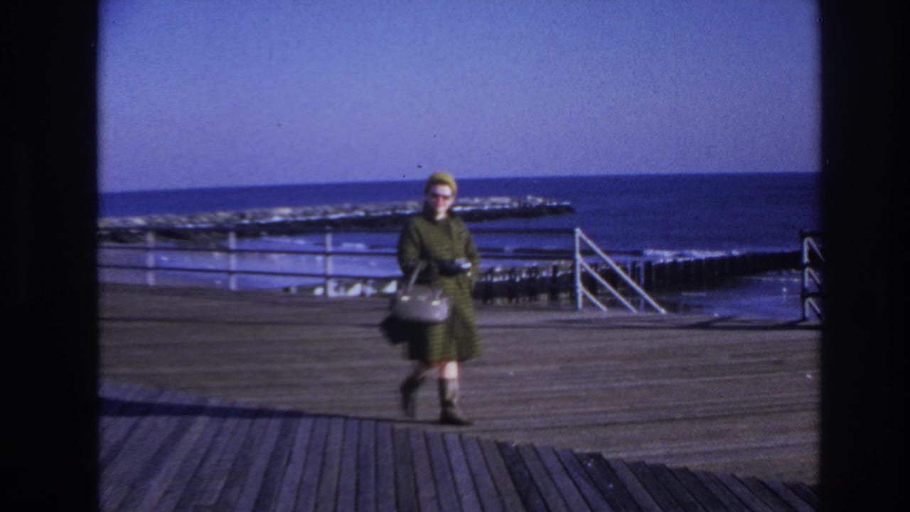 Woman in 1960s clothing walks across a Hoboken pier, carrying a light handbag as the water appears rough behind her.