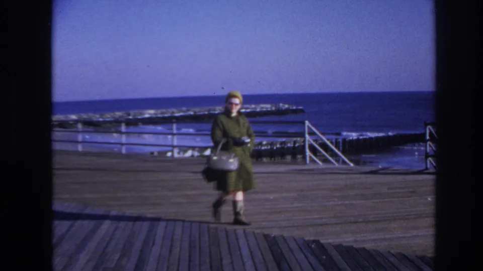 Woman in 1960s clothing walks across a Hoboken pier, carrying a light handbag as the water appears rough behind her.