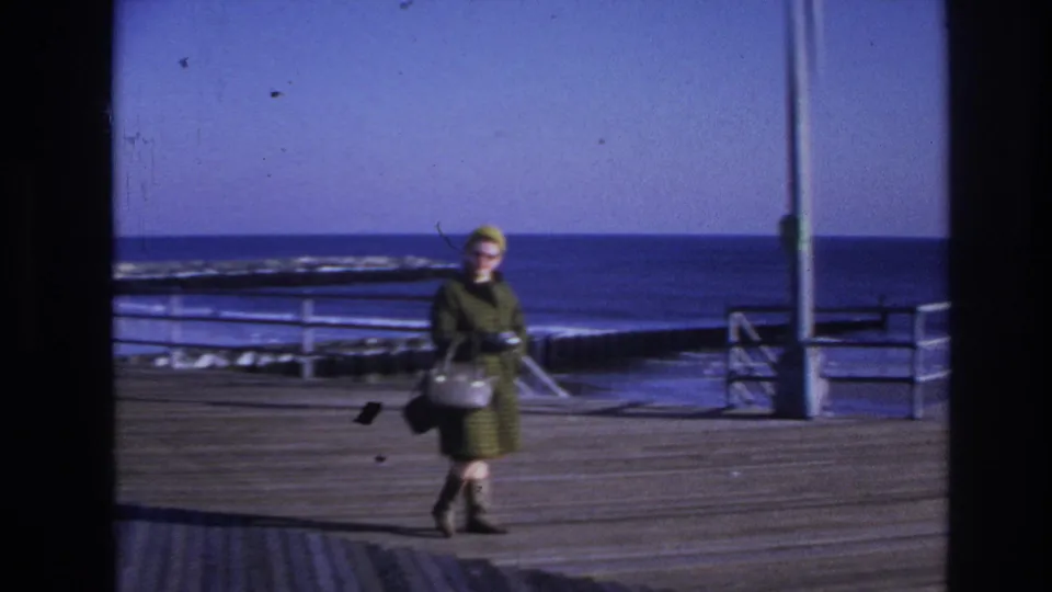 1966 Hoboken waterfront frame showing the woman walking past pier railings and a vertical pole, with choppy water beyond.