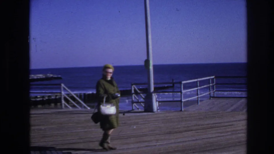 Final 1966 Hoboken pier frame with the woman in a green coat moving past railings, open water, and a clear blue sky.