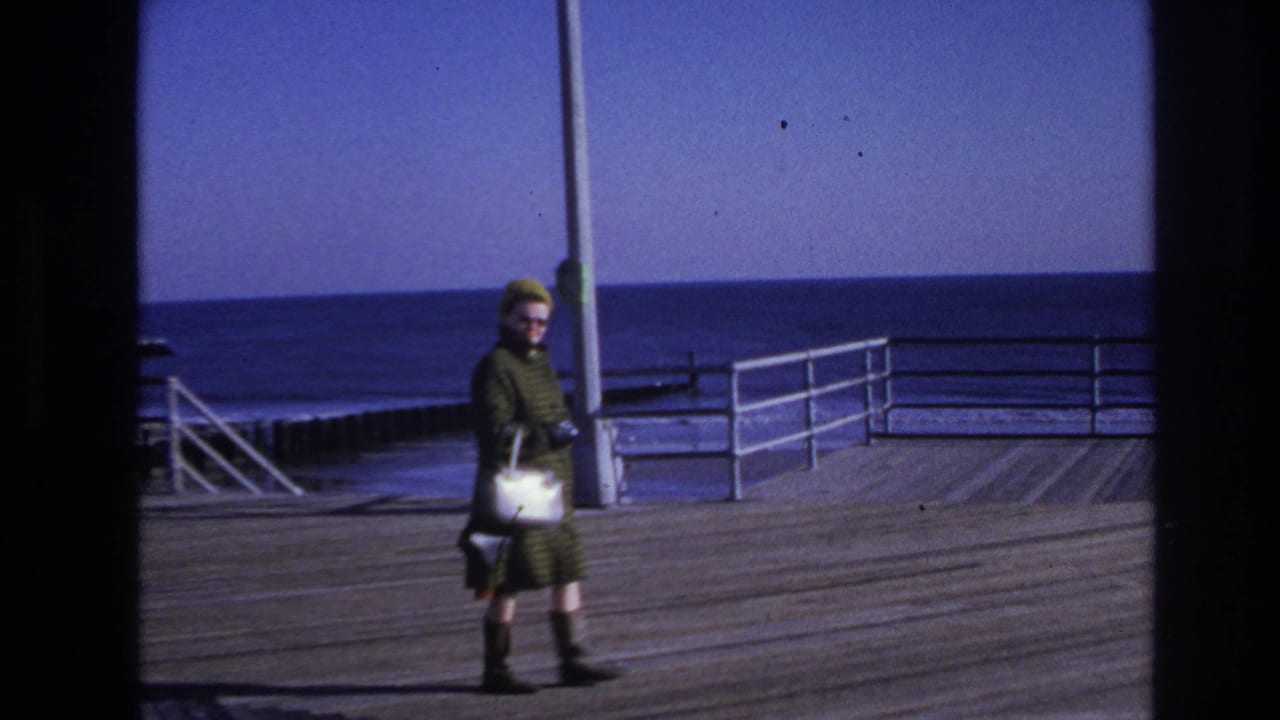 In 1966 Hoboken home-movie footage, a woman with sunglasses and a white handbag walks along a wooden boardwalk by the sea.