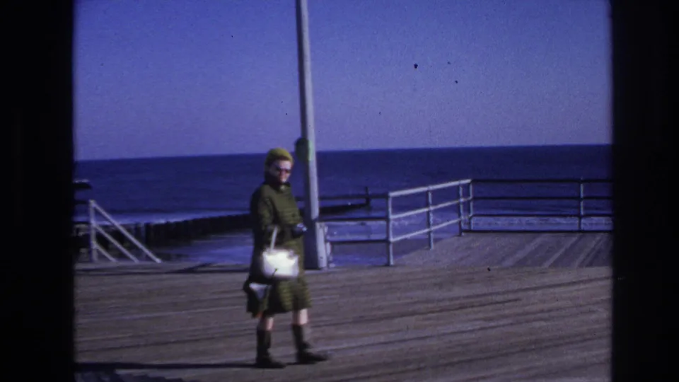 In 1966 Hoboken home-movie footage, a woman with sunglasses and a white handbag walks along a wooden boardwalk by the sea.