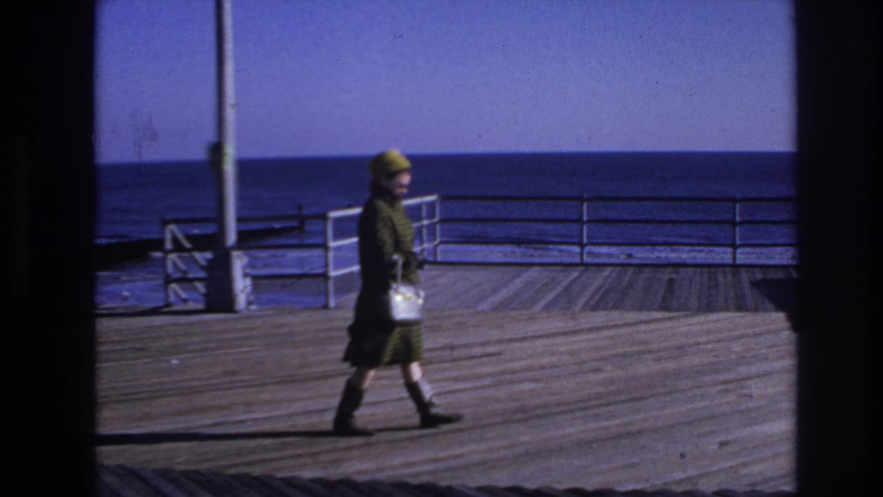 A woman in mid-century dress crosses the Hoboken boardwalk, carrying a white handbag against a blue ocean backdrop.