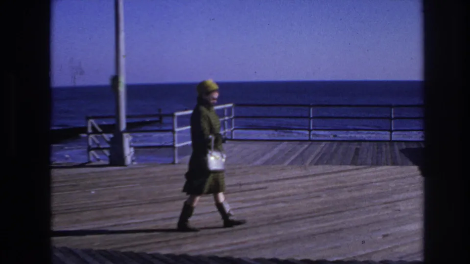 A woman in mid-century dress crosses the Hoboken boardwalk, carrying a white handbag against a blue ocean backdrop.