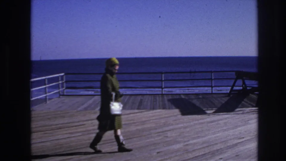The 1966 Hoboken boardwalk scene shows the woman moving past railings, with wooden planks and the ocean horizon visible.