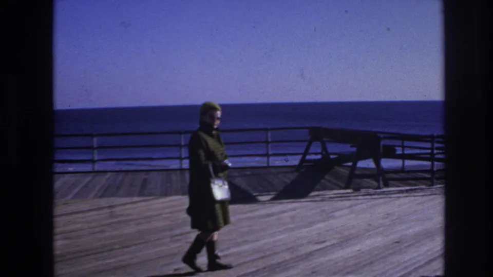 A woman with a white handbag walks through the Hoboken boardwalk frame as railings, ocean, and strong shadows define the setting.