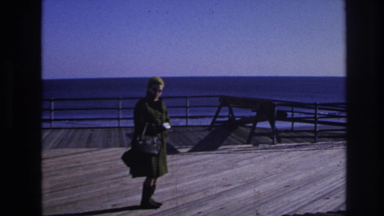 1966 Hoboken home-movie frame showing a woman in a green coat and hat standing by railing on a wooden deck near open water.