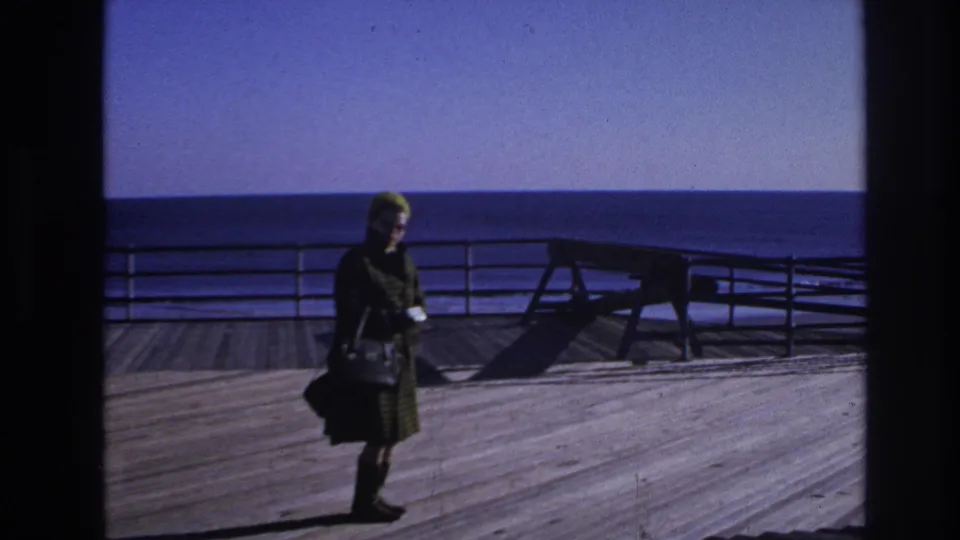 1966 Hoboken home-movie frame showing a woman in a green coat and hat standing by railing on a wooden deck near open water.
