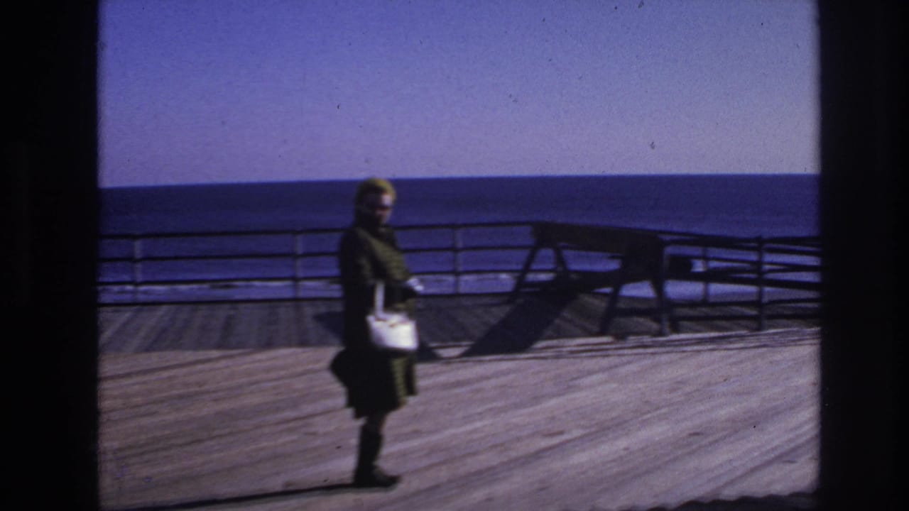 1966 Hoboken waterfront frame of a woman in dark clothing carrying a light purse as she walks along the wooden deck.