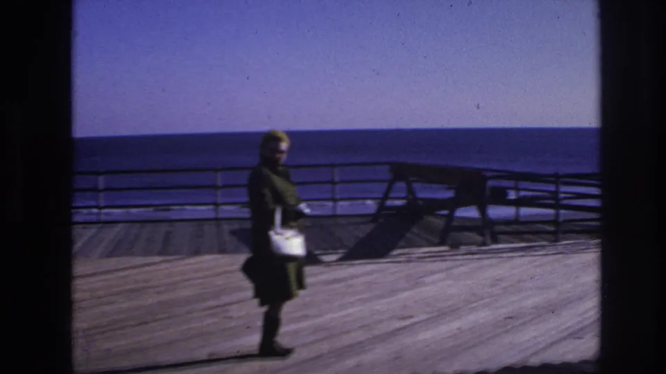 1966 Hoboken waterfront frame of a woman in dark clothing carrying a light purse as she walks along the wooden deck.