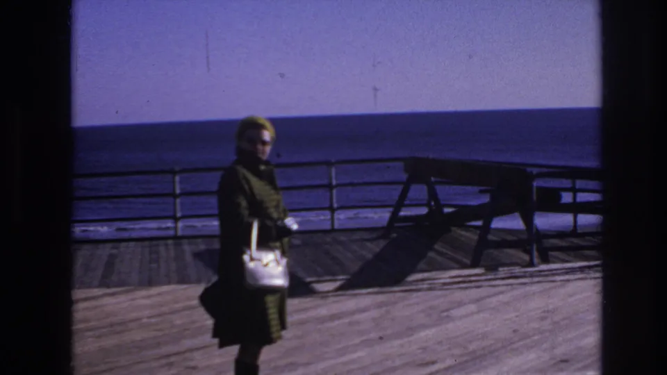 1966 Hoboken home-movie frame showing the woman nearer the camera, holding a white purse against the waterfront railing and water.