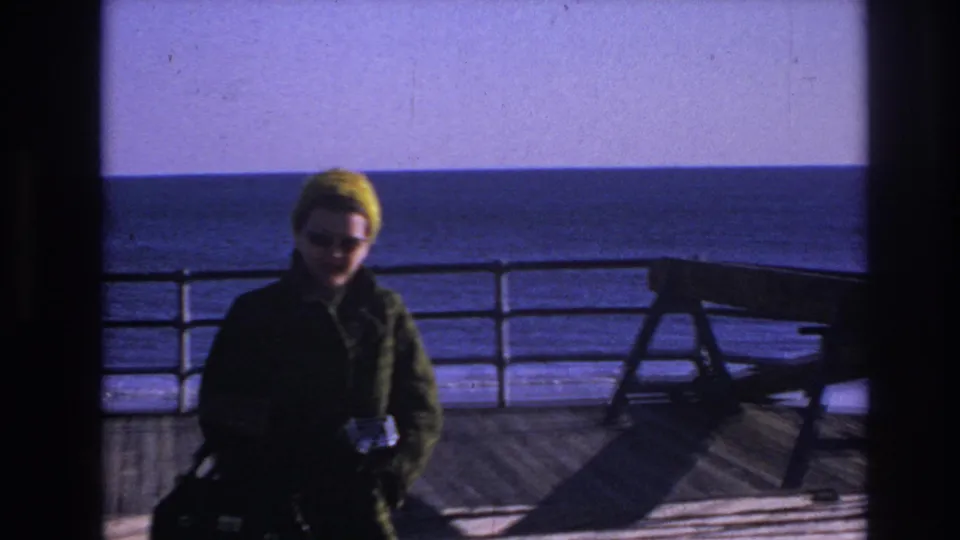 Closer 1966 Hoboken waterfront frame with the woman facing the camera on a wooden deck, bench and water visible behind her.