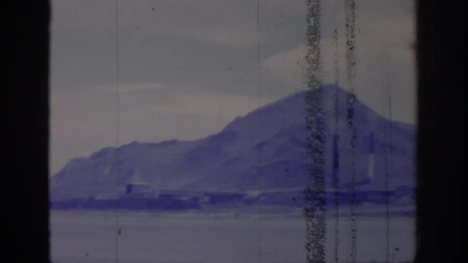 1966 8mm Sacramento-area landscape frame with mountain slope, water, shoreline forms, and dark window framing under cloudy light.