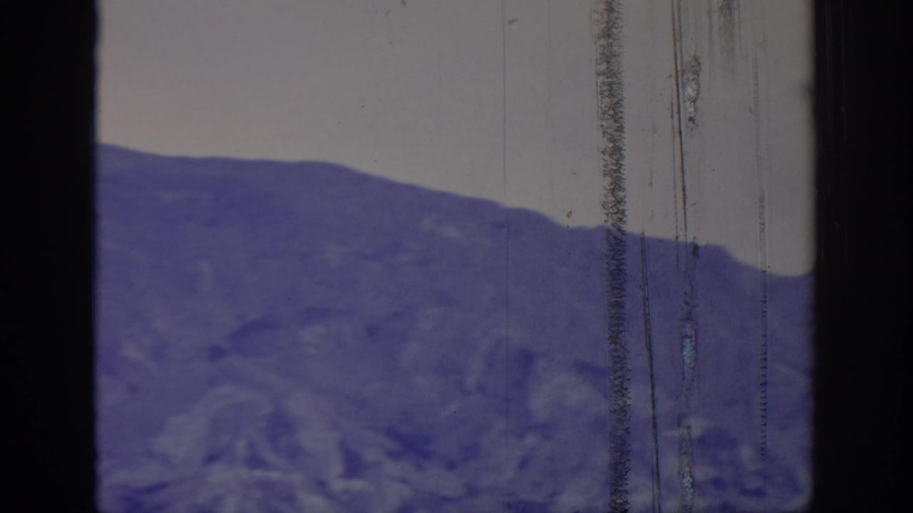 1966 8mm frame showing a distant treeless mountain ridge under a pale sky, tinted purple-blue with visible film wear.