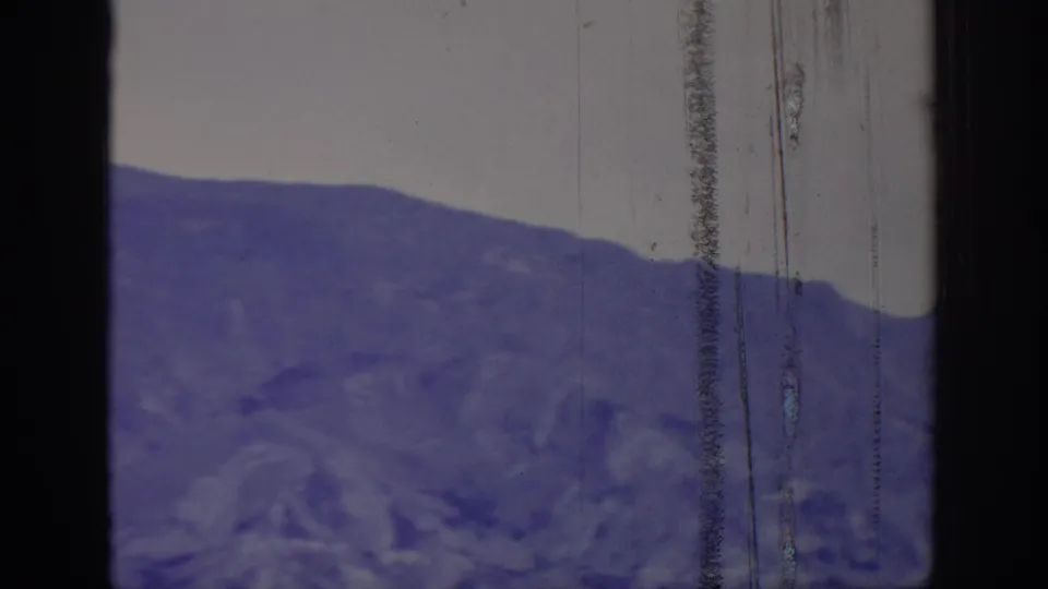 1966 8mm frame showing a distant treeless mountain ridge under a pale sky, tinted purple-blue with visible film wear.