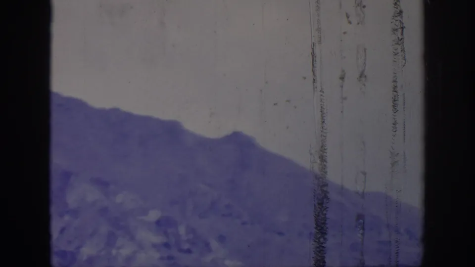 1966 8mm view of a rugged mountain slope in a purple cast, with clear sky, gate shadows, and vertical film scratches.