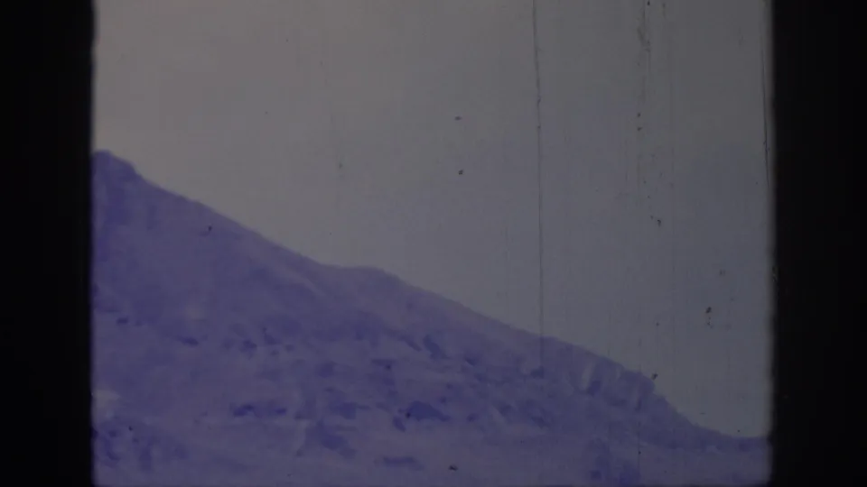 1966 8mm frame of a treeless mountain ridge seen through a worn film frame with purple tint, dust, and dark edge shadows.