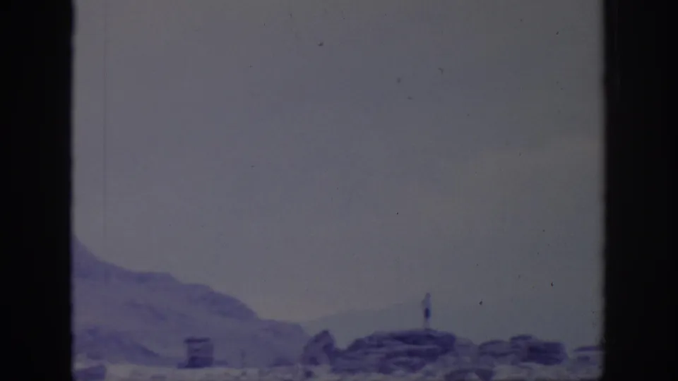 1966 Sacramento-area 8mm frame showing rocky terrain, hazy water, overcast sky, and a tiny distant figure on the ridge.