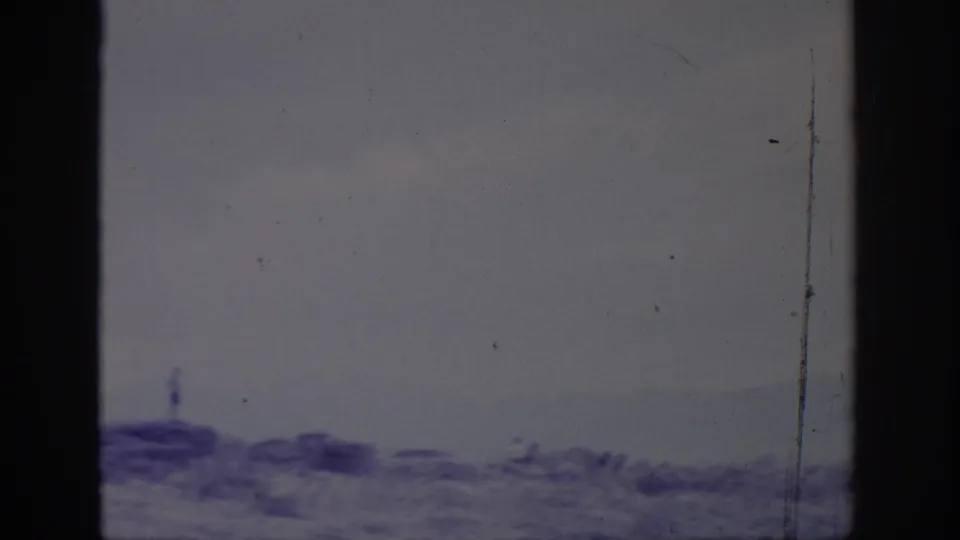 1966 Sacramento-area home-movie frame with a small figure on rocky ground overlooking pale water and distant landforms.