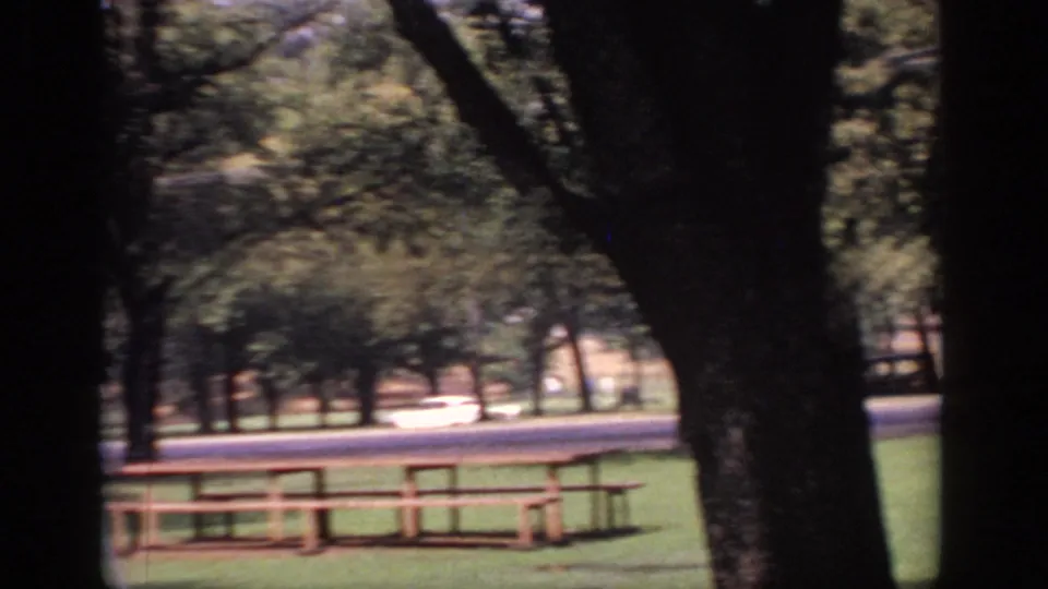 Sacramento 1966 home-movie park frame with a wooden picnic table under trees and traffic in the distance.