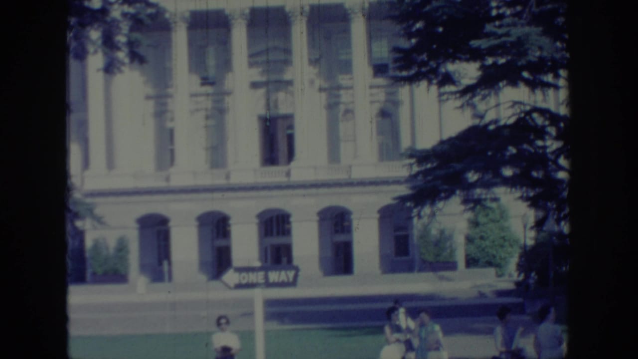 Capitol facade framed by trees with a ONE WAY sign and small pedestrians near the sidewalk.