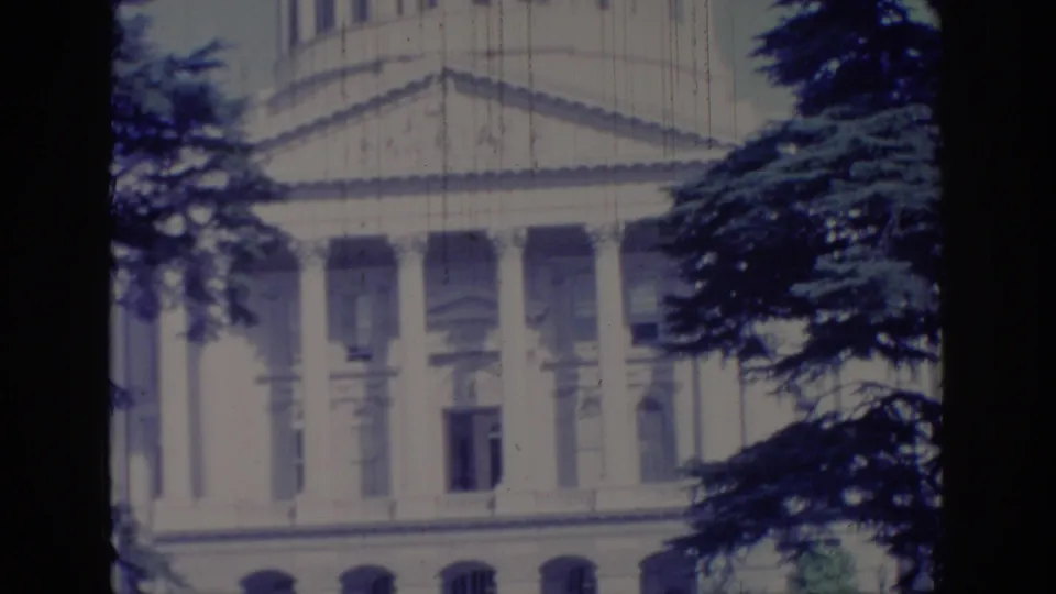 1966 Sacramento home-movie frame showing the upper California State Capitol facade, columns, and dark evergreen branches.