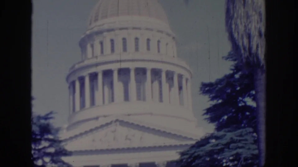 Closer 1966 home-movie view of the California State Capitol dome and facade, framed by evergreens from the lawn area.