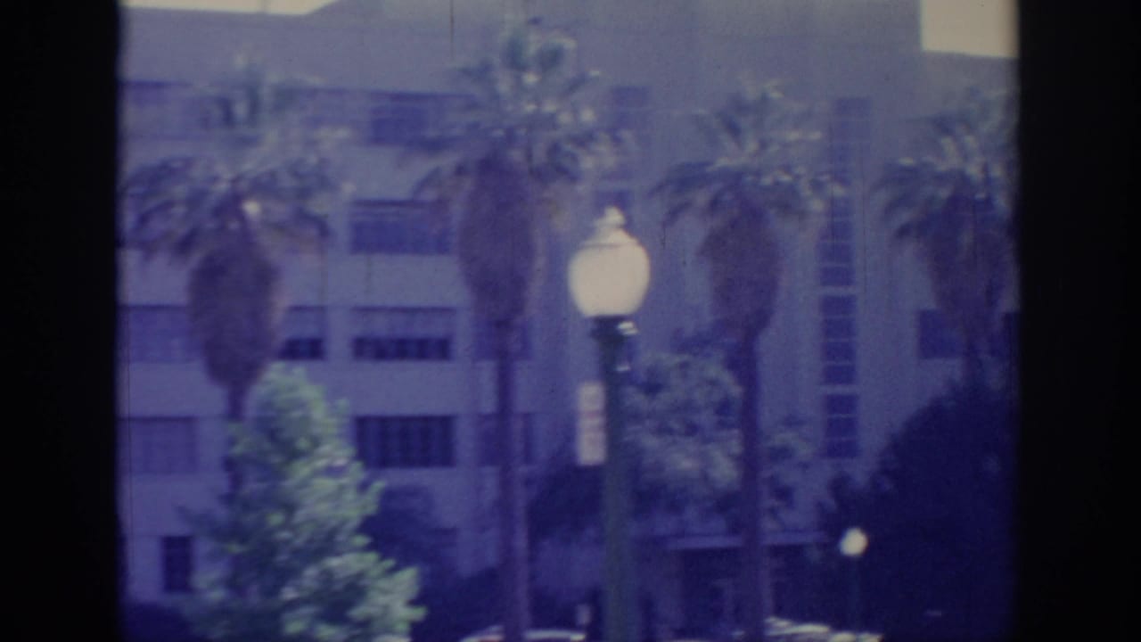1966 Sacramento home-movie frame showing a multi-story building, palms, greenery, and a white globe street lamp.